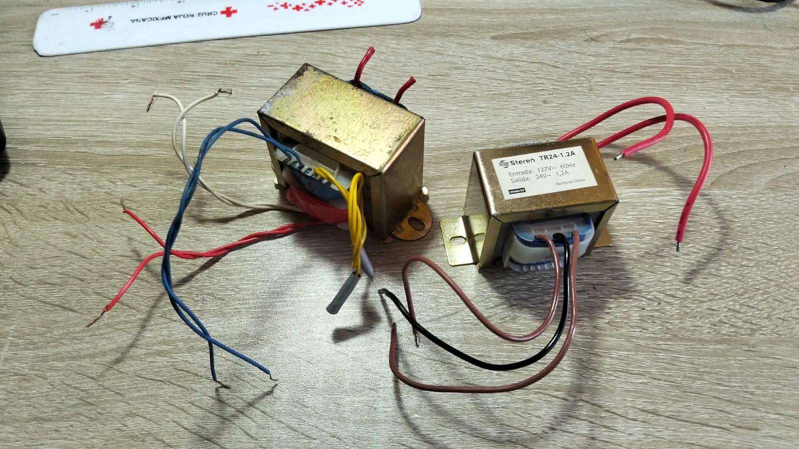 Two transformers with diferent types of outputs, displayed in a wooden background. The left one has four outputs colored yellow, white, red and blue. The right one only has a sigle output in brown, but has a central tap coded in black